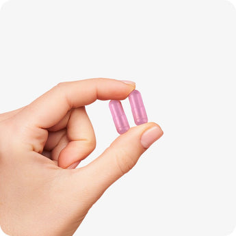 For Conceive Plus Women's Fertility Support and Ovulation Support Bundle, capsule size reference image showing a close-up of a hand gently holding two pink vegan fertility capsules between fingers, highlighting easy-to-swallow size and user-friendly design on white background.